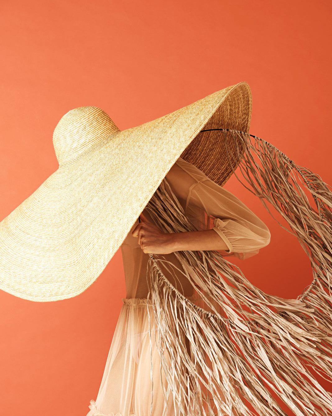 A person wearing a large beige straw hat with a wide brim, standing against an orange background.