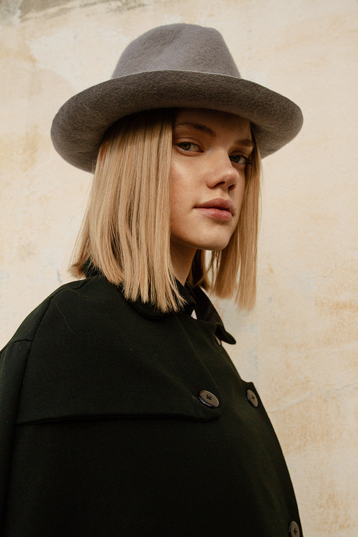 A person wearing a gray fedora hat with a curved brim and center-dent crown, paired with a black overcoat.
