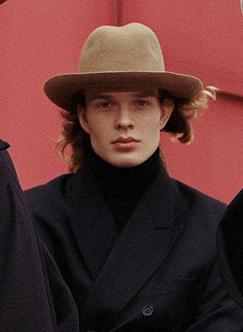 A person wearing a beige fedora hat with a curved brim and a center-dent crown, paired with black wool overcoat.