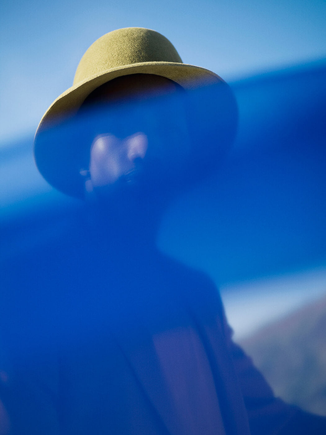 A person wearing a khaki-colored fedora hat with a center-dent crown and a sloping brim, standing in front of a blue chiffon fabric.
