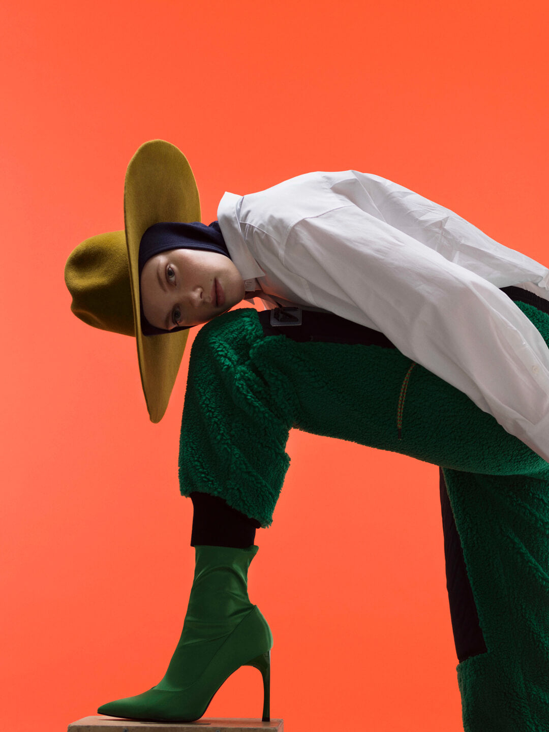 A person wearing a beige-yellow flat-brimmed fedora hat and green patterned pants, with a white shirt and green boots.
