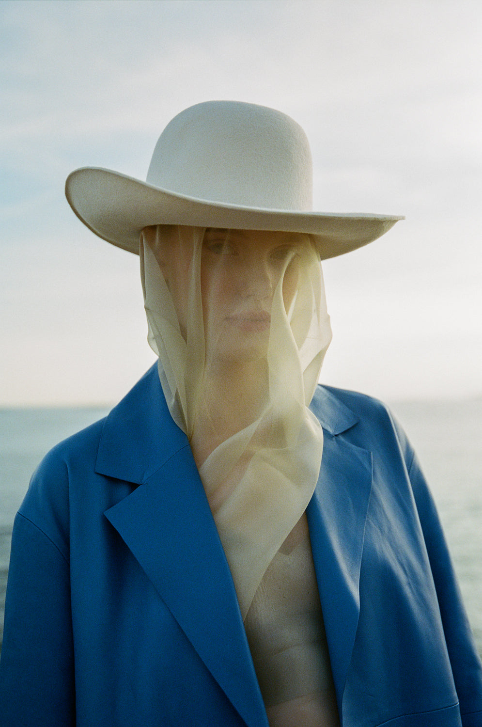 A person wearing a white cowboy hat with an upturned brim and a softly indented crown, outdoors with a blue coat and a scarf covering the lower face.