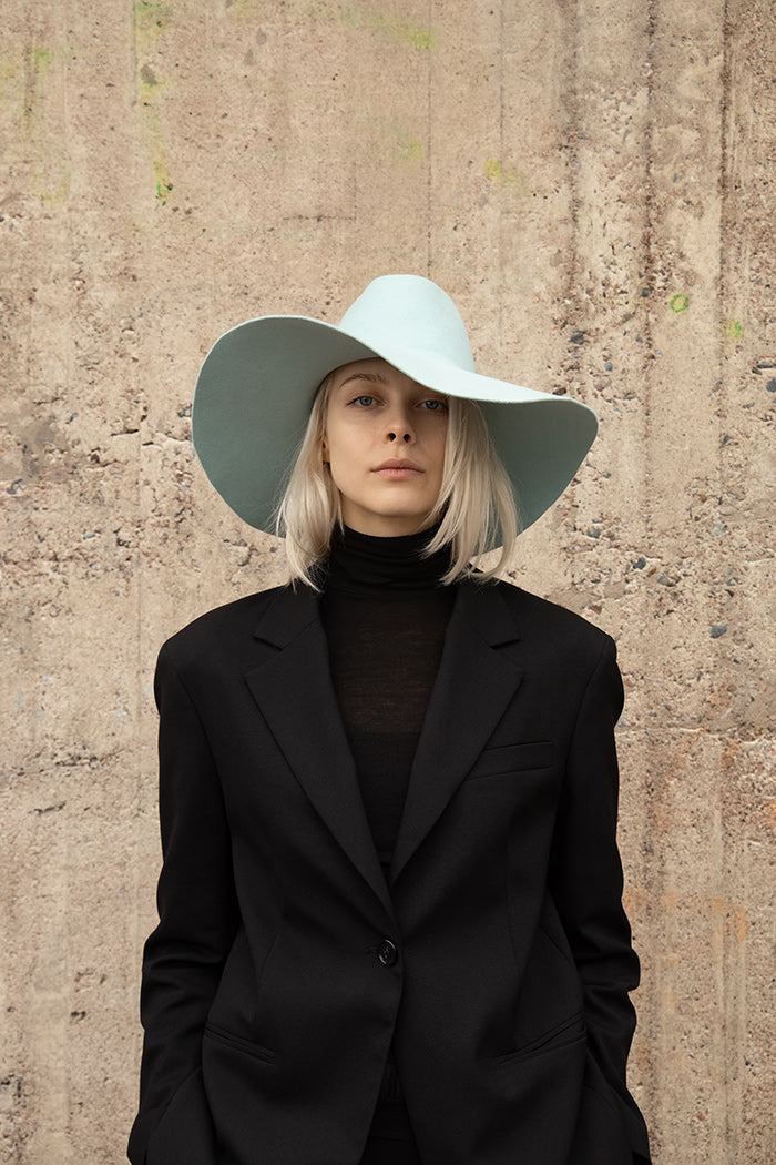 A person wearing a light blue felt hat with a round flat brim and a round crown paired with black blazer and turtle-neck knit blouse in front of beige wall.